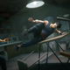 A screenshot from Sifu depicting the protagonist jumping over a table to kick two people in a small, dark room.
